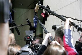 Fernseh- und Hörfunkjournalisten mit Kamera und Mikrofonen scharen sich um einen Mann, der ein Interview gibt. 