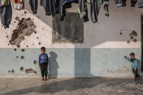 Syrische Kinder spielen vor einem Haus, dessen Wand durch Einschusslöcher beschädigt ist.